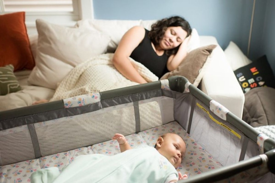 Mom naps on couch while infant less than 1 year old lays in infant safe crib.